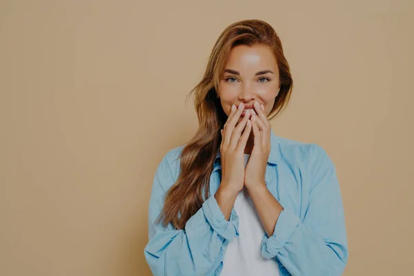 Happy female model with cheerful expression covers her lips with hands, smiling shyly at camera, wears casual white tshirt and blue shirt, isolated over beige background. Human facial expressions