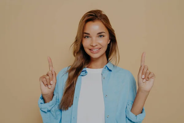 Charming caucasian young female pointing forefingers upwards at copy space, having great idea and expressing joy and happiness while standing isolated on beige background. Advertisement and promotion