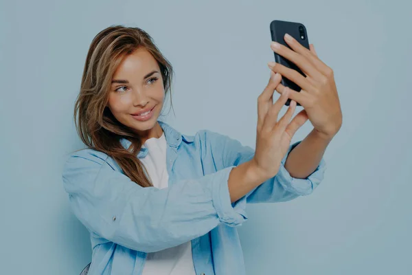 Attractive female blogger holding modern smartphone and taking selfie, broadcasting or filming video for social media, positively smiling at camera while posing on blue wall. People and technologies