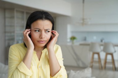 Girl is irritated with noise and closes her ears. Conceptual portrait of young emotional woman. Advertising banner mockup. Handsome european girl in living room at home. Modern interior of apartment.