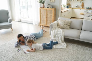 Avrupalı anne çocuğa resim çizmeyi öğretiyor. Aile hafta sonu evde. Genç anne ve küçük kız birlikte yerde yatıyor ve renkli kalemlerle resim yapıyorlar. Kadın ve çocuğun evde boş vakitleri var..