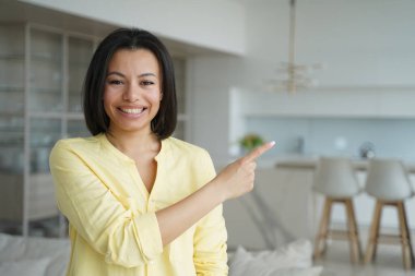 Smiling female realtor homeowner or tenant pointing with finger at copy space aside standing in new home. Happy positive woman recommend shopping offer, advertise service, looking at camera.