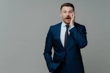 İnanılmaz bir haber. Takım elbiseli genç işadamı şaşırmış yüz ifadesiyle kameraya bakıyor ve gri arka planda dururken ağzını açıyor. İş adamları ve duygu kavramı