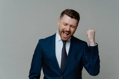 Olumlu iş adamı sevinç çığlıklarından yumruğunu sıkıyor. Gri arka planda izole edilmiş başarılı iş anlaşmasının ardından resmi olarak tatmin olan başarıyı kutluyor. Tebrikler. Başardım.