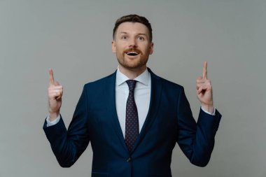 Şok olmuş takım elbiseli iş adamı parmaklarıyla işaret ediyor ve gri arka planda dururken ağzı açık heyecanlanıyor. Erkek girişimci harika bir yenilikçi fikre sahip.