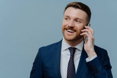 Takım elbiseli başarılı işadamı iş görüşmeleri yapıyor ve gülümsüyor. Açık mavi arka planda tek başına duruyor. Erkek girişimci cep telefonuyla konuşmaktan hoşlanıyor.
