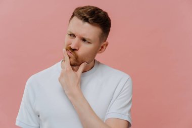 Tıraşsız, beyaz gömlekli düşünceli genç bir adam. Pembe arka planda tek başına dururken kafası karışmış bir yüz ifadesi olan bir şey düşünüyor. Tereddüt