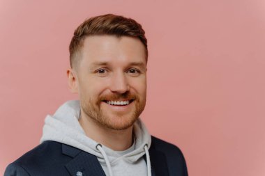 Pembe arka planda izole bir şekilde dikilirken kameraya gülümseyen sakallı neşeli genç adamın portresi. Yüzünde olumlu ifade olan kızıl saçlı yakışıklı adam.