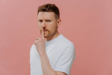 Sır olarak sakla. Tıraşsız, beyaz tişörtlü, sessiz jest yapan gizemli yüz ifadesiyle kameraya bakan pembe stüdyo arka planında duran genç bir adam.