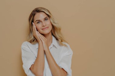 Sıradan beyaz tişörtlü romantik sarışın bayan ellerini yüzüne yakın tutuyor ve kameraya nazikçe gülümsüyor, bej arka planda fotokopi alanı ile poz veriyor. İnsan yüzü ifadeleri