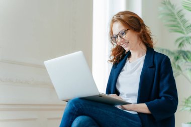 Profesyonel kadın işveren çalışma işlemi sahiptir, çapraz bacakları dizüstü bilgisayar aygıtla oturur, çevrimiçi sohbetler, olumlu görünüme sahip, kırmızı kıvırcık saçlı, şık zarif giysiler giyiyor. İnsan ve teknoloji