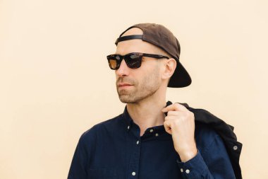 Siyah şapkalı, şık gözlüklü, şık tişörtlü, bej arka planda izole edilmiş, düşünceli bir adam. Moda dergisi için çekici erkek pozları. İnsanlar ve tarz kavramı