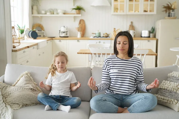 Mother and little daughter practice yoga, family meditation, sitting on sofa at home. Calm mom or babysitter and kid girl relax together sit in lotus position on couch. Healthy lifestyle, parenting.