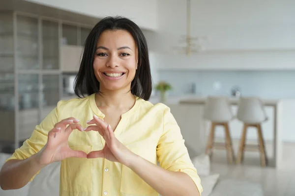 Friendly loving woman feel grateful show heart gesture, sign of love, support and care. Smiling female blogger volunteer express appreciation, ask audience to support charities, standing at home.