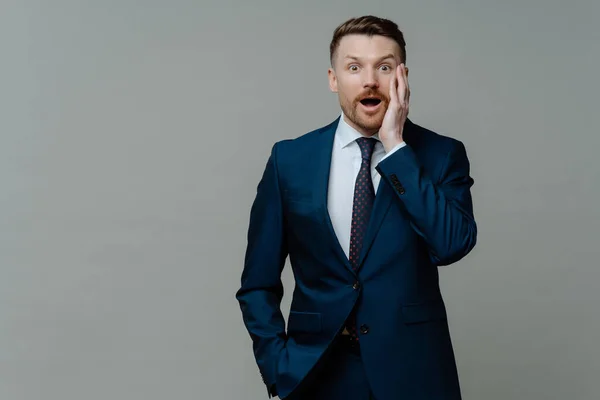 stock image Unbelievable news. Young shocked businessman in suit looking at camera with surprised face expression and opened mouth while standing against grey background. Business people and emotions concept