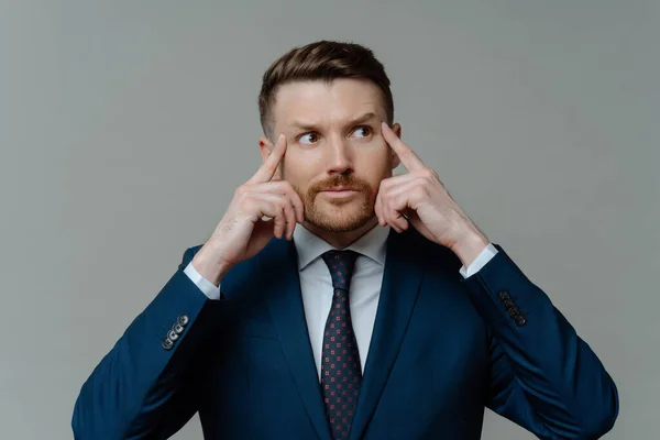 stock image Indoor shot of motivated thoughtful bearded businessman keeps fingers on temples looks away gets ready for conference dressed in formal clothes isolated over grey background has hard working day