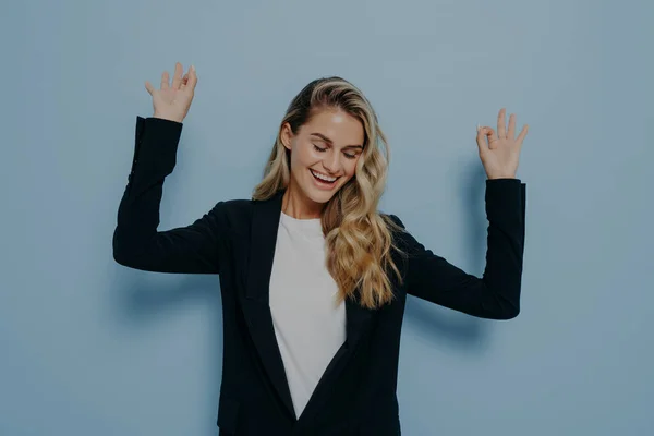 Portrait of carefree happy woman with blonde dyed hair raising her arms and dancing with closed eyes and smile on face while standing isolated against blue background. Happiness concept