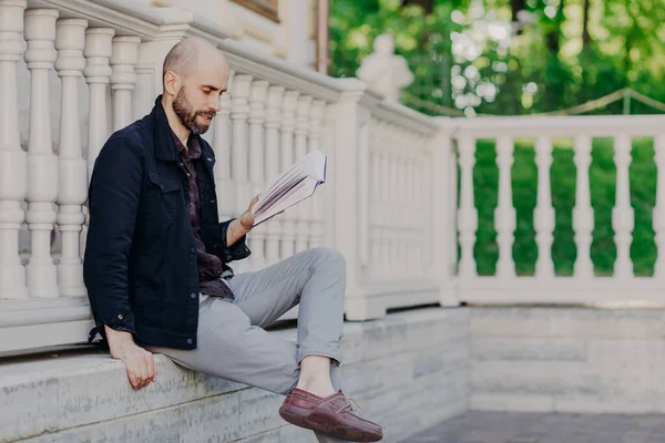 People, learning and education. Bearded young male student reads book as prepares for classes, develops himeself and improves knowledge in particular sphere, poses outdoor alone, being serious
