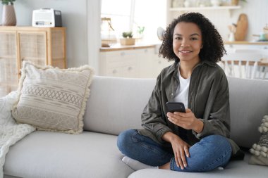 Young hispanic woman is browsing social media and smiling. Curly african american girl has fun with phone at home. Chatting with friends remotely. Lazy weekend morning. Online book reading.