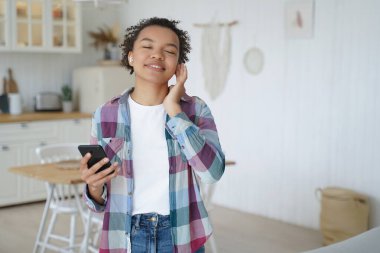 Music listening in airpods on mobile phone. Teenage girl enjoying music in wireless earphones. Afro young woman relaxing at home. Having fun and recreation alone. Music service website or app concept.