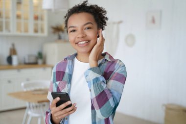 Kablosuz kulaklıkla kablosuz müzik dinlemekten hoşlanan mutlu afro kız. Cep telefonundan hava podcasıyla müzik dinletiliyor. Evde dinlenme ve dinlenme. Çekici genç bayan eğleniyor..