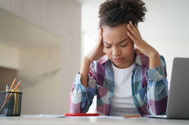 Tired african american teenage schoolgirl suffer headache while doing homework at laptop at home. Exhausted, frustrated teenager girl worried about educational problem or bullying. Difficult elearning