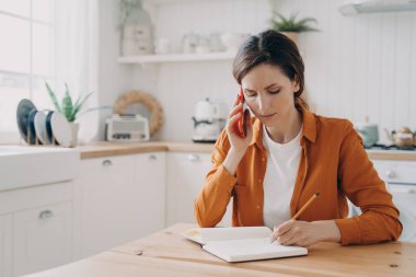 European girl is sitting at the kitchen, taking notes and talking on phone. Tired young woman is remote employee. Secretary, office manager or customer support assistant. Distance work concept.
