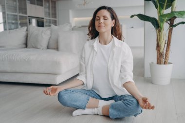 Kafkasyalı genç bir kadın Lotus Asana 'da oturuyor. Kız evde yoga ve meditasyon yapıyor. Sabah jimnastiği. Uyum ve zihin rahatlatma kavramı. Oturma odasının İskandinav iç mekanı.