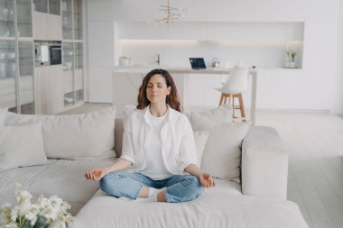 Genç İspanyol kadın evdeki kanepede yoga ve meditasyon yapıyor. Duruş egzersizi, Lotus Asana. Sabah jimnastiği, sağlıklı yaşam tarzı. Konsantrasyon ve zen bulma kavramı.