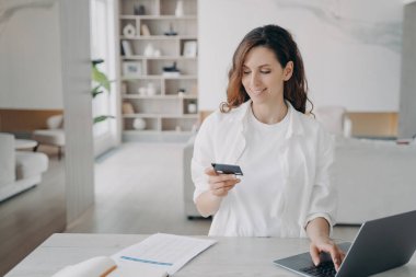 Happy spanish girl is holding credit card in front of pc. Young pretty woman is going to buy through internet. Girl is booking or purchasing online. E-commerce and financial transaction concept.