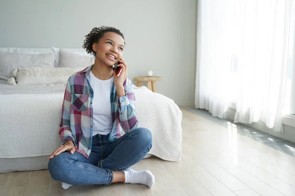 Morning phone call. Young african american woman is talking to friend on smartphone in bedroom. Cute spanish teenage girl in casual wear is phone addict. Communication and relaxation.
