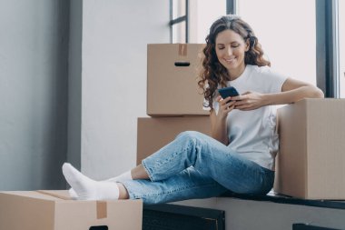 Happy spanish woman is sitting on windowsill of new home. Girl is unpacking boxes. Young lady is texting on phone and smiling. Joy of relocation concept.