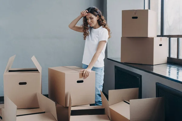 Puzzled woman unpacking cardboard boxes. Tired girl in new apartment. Lady touches forehead with hand, gesture of tired person. Packing things, shipping service and relocation concept.