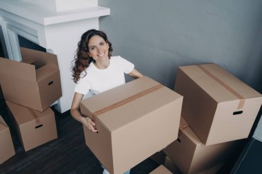 Attractive european girl in white t-shirt is leaving house. Lady is packing cardboard boxes and carrying. Cheerful young woman is going to buy apartment. Relocation and independence concept.