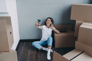 Happy young spanish woman in jeans and white t-shirt unpacking boxes. Girl having video call on phone and sitting on the floor. Enjoyment of relocation and distance friendship concept.