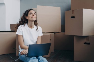 Hopeful hispanic woman sitting among cardboard boxes and holding pc. Lady is in waiting for shipping service and chatting online. Romantic relationship and relocation concept.