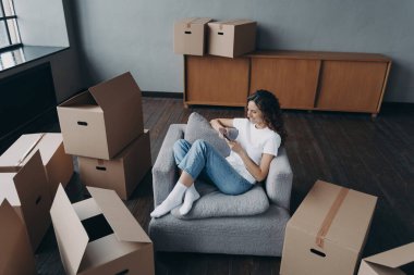 Girl having rest in new apartment and clicks phone. Successful independent spanish woman is moving. Living room interior and cardboard boxes. Mortgage loan and dream house purchase concept.