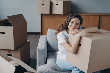 Hispanic girl relocates to new home. Lady is sitting in armchair among packed cardboard boxes. Woman is dreaming leaned to box. Happy young woman moves alone. Independence and success concept.