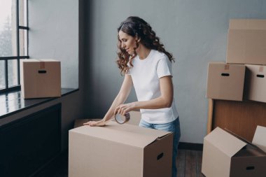 Curly spanish girl is ready for relocation. Lady is wrapping cardboard boxes with packing tape. Happy young woman purchasing or selling real estate and going to send things as cargo.
