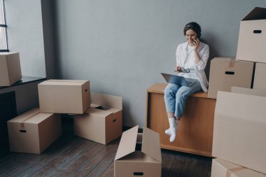 Happy homeowner talking on phone. Hispanic girl is sitting among carton boxes and working on pc. Attractive young business woman moves. Mortgage loan and real estate purchase.