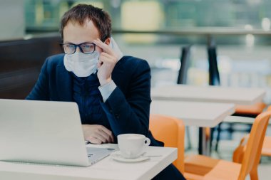 Tıbbi maskeli ve gözlüklü bir adam, modern dizüstü bilgisayarın önünde oturur, rahat kafeteryada poz verir, Coronavirus hakkında haberler okur, sağlıkla ilgilenir, kahve içer. Salgın Karantina