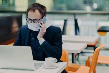 Ciddi meşgul bir işçi karantinada çalışıyor, dizüstü bilgisayardan bilgi okuyor, koruyucu tıbbi maske takıyor, cep telefonuyla birilerini arıyor, salgın virüs enfeksiyonu sırasında sağlığa önem veriyor.