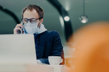İşadamı kafeteryada uzaktan çalışıyor, dizüstü bilgisayar ekranında yoğunlaşıyor, telefonla konuşuyor, grip veya koronavirüs epidemisine karşı korunmak için yüzüne tıbbi maske takıyor. Akıllı çalışma