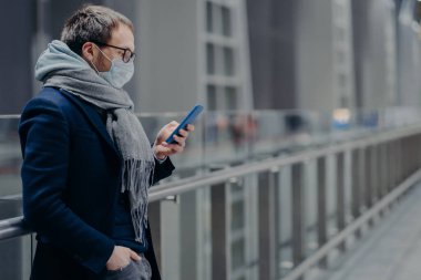 Ciddi Avrupalı adam akıllı telefona bakar, internetten haber okur, mesaj gönderir, virüsten korunur, tıbbi maske takar, koronavirüs korumasını önemser. Virüs salgını, Covid-19