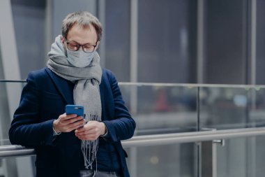 Gözlüklü adamın yatay görüntüsü modern cep telefonu kullanıyor, Coronavirus salgını sırasında maske takıyor, büyük bir alışveriş merkezinde gribe karşı korunuyor. Grip ve korona mevsimi. Sağlık hizmeti kavramı