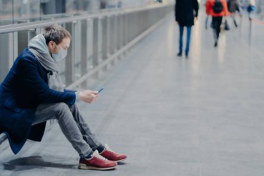 Adamın yatay görüntüsü modern cep telefonu, salgın ve koronavirüs sırasında alışveriş merkezinde, koruyucu maske takıyor, günlük kıyafetlerini giyiyor, mobil uygulamaları kontrol ediyor. Sağlık hizmeti kavramı