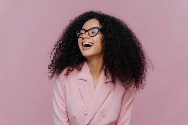 Neşeli koyu renk tenli kadın menajer kıvırcık saçlı, mutluluktan kahkahalar atan, zarif bir kostüm giyen, şeffaf gözlük takan, odaklanmış, mor arka planda poz veren. Duygular konsepti
