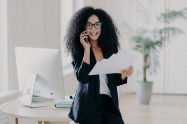 Afro saç stili olan zeki bir kadın, elinde kağıt belgeler var, bilgileri kontrol ediyor, cep telefonuyla iş ortağını arıyor, gelecekteki stratejisini analiz ediyor, bankacılık müdürüyle konuşuyor, siyah takım elbiseli.