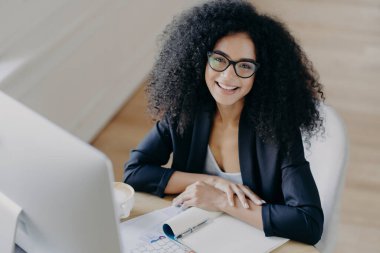 Afro-Amerikan iş kadınlarının mutlu bakışları hoş bir şekilde gülümsüyor, bilgiyi not alıyor, notlar alıyor, şeffaf gözlük takıyor, resmi kıyafetler giyiyor, bilgisayar monitörü önde tek başına oturuyor.