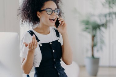 Mutlu kıvırcık gencin kırpılmış görüntüsü cep telefonuyla aktif bir konuşma yapıyor, gözlük takıyor ve sarafan takıyor, kolunu kaldırıyor, telefon görüşmeleri sırasında el kol hareketi yapıyor, neşeli bir ifade takınıyor.
